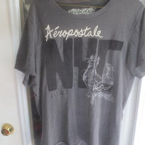 May include: A grey short-sleeved t-shirt featuring the white text Aéropostale at the neckline and large black letters NYC across the chest. A white line-art graphic of a bird is layered over the letters, with the number 1987 printed below.