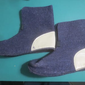May include: A pair of dark blue felted boots featuring a high-top silhouette and a smooth, cream-colored synthetic heel patch. The boots have a simple, unadorned design with a rounded toe and a flat sole, showcasing a textured fabric construction.