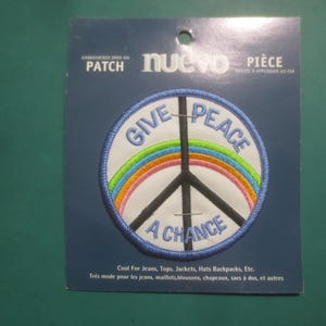 Peut inclure: Écusson brodé thermocollant avec un symbole de paix. Le patch présente un fond blanc avec un symbole de paix noir, un arc-en-ciel et les mots "GIVE PEACE A CHANCE" en bleu. Le patch est sur une carte bleue.