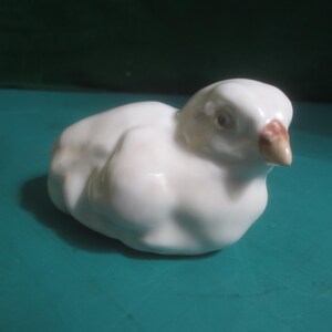 May include: A small white ceramic figurine of a resting bird. The bird has a smooth, glossy finish with a rounded body shape, a small light brown beak, and a single visible dark eye. The sculpture is depicted in a seated or nesting position.