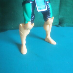 Op de afbeelding: Het onderlichaam van een plastic actiefiguur met turquoise en zwarte dijbeenbescherming en een centrale turquoise tabard. De onderbenen en voeten zijn gegoten in een effen lichtbeige kleur, met duidelijk zichtbare kuitspieren en voeten.