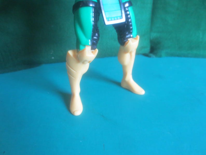 Op de afbeelding: Het onderlichaam van een plastic actiefiguur met turquoise en zwarte dijbeenbescherming en een centrale turquoise tabard. De onderbenen en voeten zijn gegoten in een effen lichtbeige kleur, met duidelijk zichtbare kuitspieren en voeten.