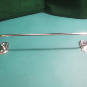 May include: A chrome-finished towel bar with a straight horizontal bar and curved ends. The bar is mounted on two rectangular brackets with screw holes. The background is a teal surface with a green fabric.