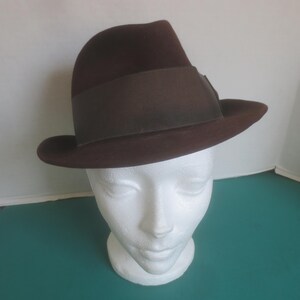Peut inclure: Un chapeau fedora marron avec un ruban marron foncé. Le chapeau est présenté sur une tête de mannequin blanche. Le chapeau a un design classique avec un bord légèrement incurvé et une couronne pincée. Le chapeau est en feutre.