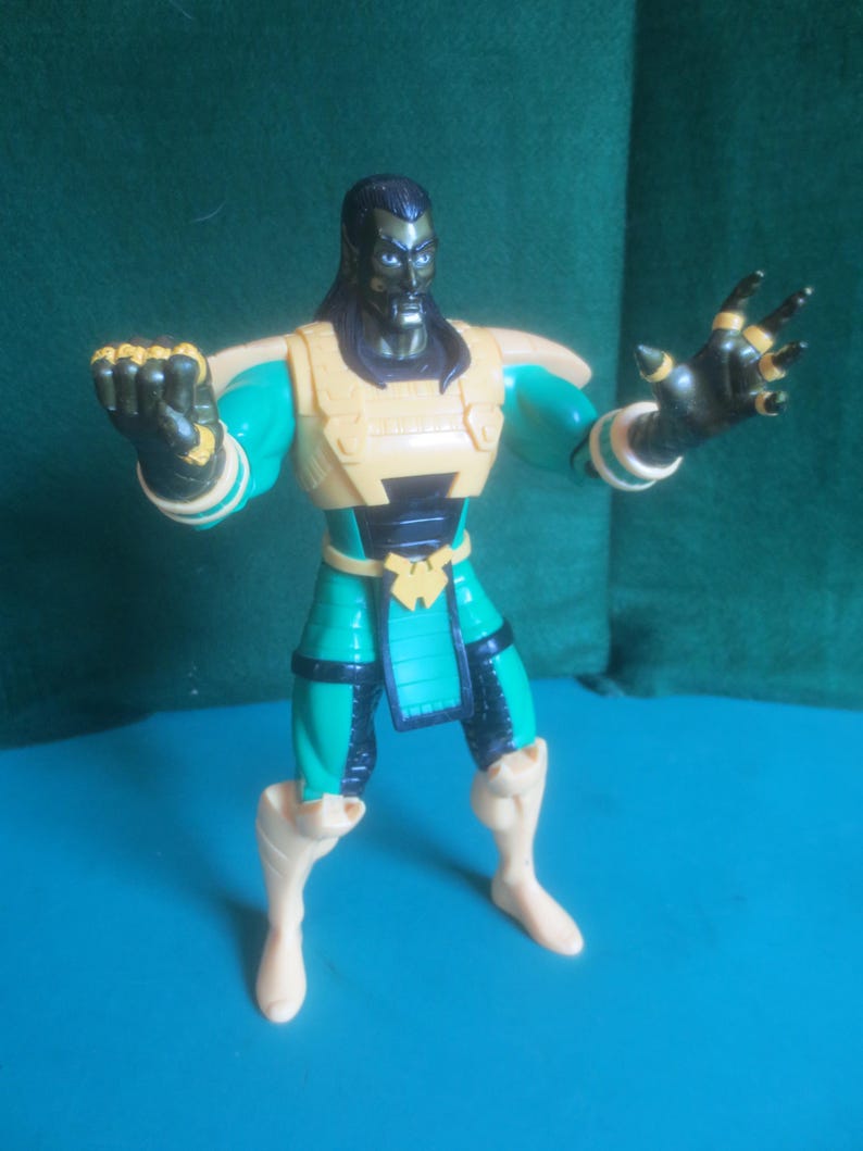 Op de afbeelding: Een plastic actiefiguur van een donkerhuidig personage met lang zwart haar, gekleed in een groenblauw en zwart pak met een gele borstplaat, schouderstukken en riem. De figuur heeft &eacute;&eacute;n vuist gebald en de andere hand is open.