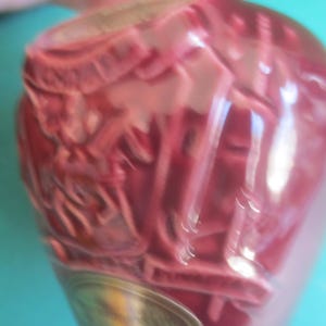 May include: A close-up of a pink, decorative glass bottle with an ornate design. The bottle has a silver-colored base and is set against a turquoise background. The bottle's surface is textured.