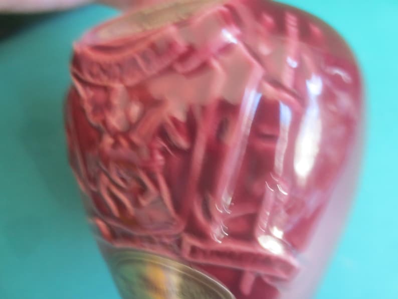May include: A close-up of a pink, decorative glass bottle with an ornate design. The bottle has a silver-colored base and is set against a turquoise background. The bottle's surface is textured.