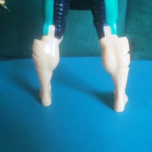 Op de afbeelding: Een onderste deel van een actiefiguur van kunststof met beige benen, gedetailleerde kuitspieren en donkerblauwe en blauwgroene dijbeenpantsers. Het figuur is vanaf de taille naar beneden afgebeeld, met scharnierende gewrichten bij de heupen en knie&euml;n en een gladde, glanzende afwerking op de gegoten kunststof onderdelen.
