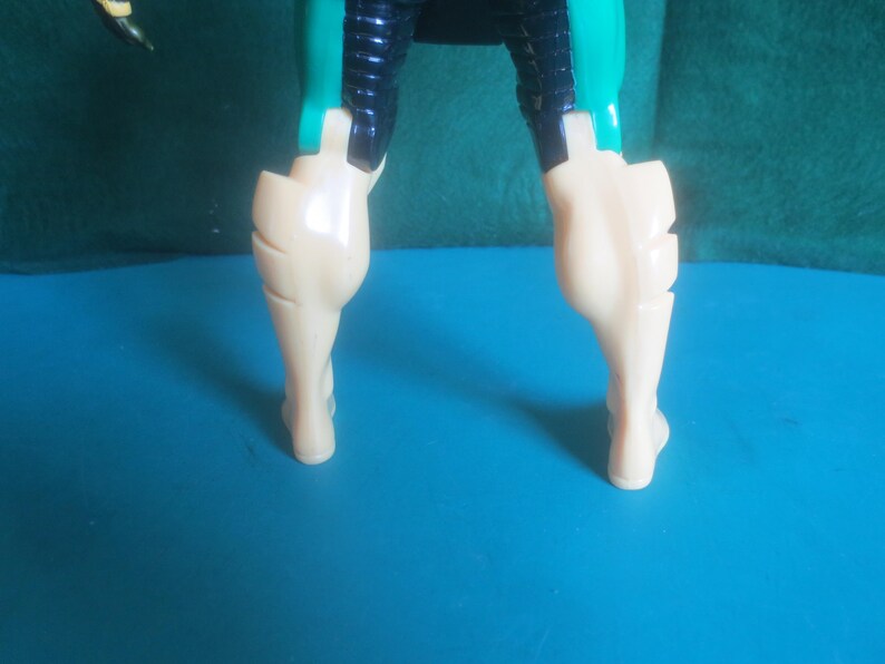 Op de afbeelding: Een onderste deel van een actiefiguur van kunststof met beige benen, gedetailleerde kuitspieren en donkerblauwe en blauwgroene dijbeenpantsers. Het figuur is vanaf de taille naar beneden afgebeeld, met scharnierende gewrichten bij de heupen en knie&euml;n en een gladde, glanzende afwerking op de gegoten kunststof onderdelen.