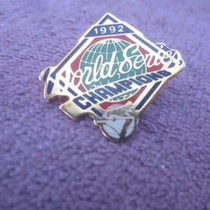 Può includere: Spilla da baseball dorata a forma di diamante, con la scritta "World Series Champions" e l'anno "1992". La spilla presenta un disegno del globo e dettagli rossi, bianchi e blu. Su sfondo viola.