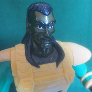 Op de afbeelding: Een plastic actiefiguur met een donkergroene huid, zwart haar en een snor. Het figuur draagt een gele borstplaat met schoudervullingen over een donkergroen shirt. De armen zijn groen met gele polsmanchetten en hebben een gedetailleerd ontwerp.