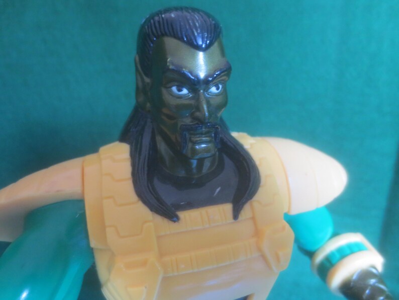 Op de afbeelding: Een plastic actiefiguur met een donkergroene huid, zwart haar en een snor. Het figuur draagt een gele borstplaat met schoudervullingen over een donkergroen shirt. De armen zijn groen met gele polsmanchetten en hebben een gedetailleerd ontwerp.