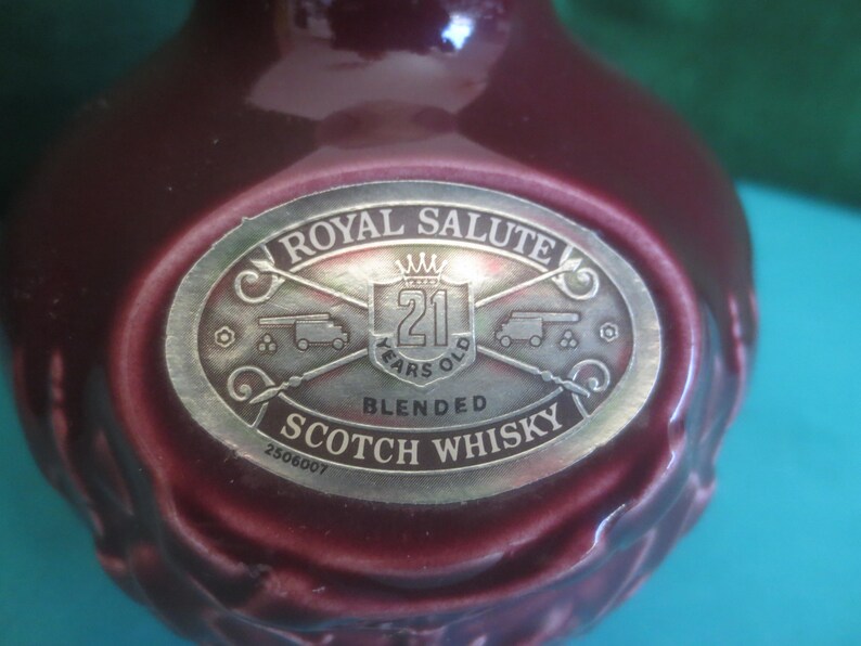 May include: A maroon-colored bottle with a silver label that reads "Royal Salute 21 Years Old Blended Scotch Whisky." The label features a crown, cannons, and the text "2506007."