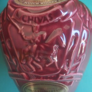 May include: A burgundy-colored Chivas Regal blended Scotch whisky bottle. The bottle has a textured surface with embossed details and the brand name prominently displayed. The bottle has a decorative design with a gold label.