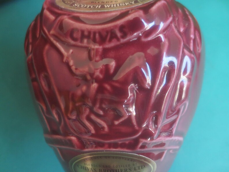 May include: A burgundy-colored Chivas Regal blended Scotch whisky bottle. The bottle has a textured surface with embossed details and the brand name prominently displayed. The bottle has a decorative design with a gold label.