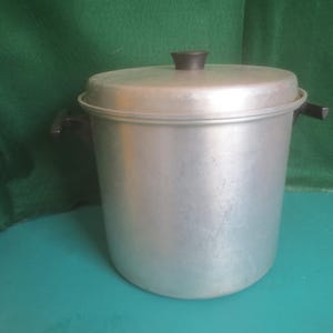 May include: A large, cylindrical aluminum stockpot with a matching lid. The pot features two dark brown, heat-resistant side handles and a matching dark brown knob centered on the lid. The exterior surface has a matte, brushed metal finish.