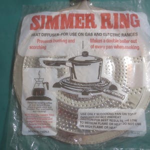 May include: A silver simmer ring heat diffuser for use on gas and electric ranges. The packaging features the text "SIMMER RING" in red, with illustrations of a pot and coffee maker. It prevents burning and scorching.