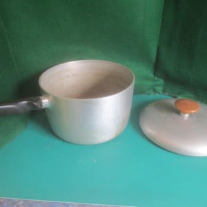 Peut inclure: Une casserole en aluminium de couleur argentée avec une poignée noire et un couvercle assorti doté d'un petit bouton rond marron. L'intérieur de la casserole présente des traces d'utilisation avec une décoloration marron clair au fond.