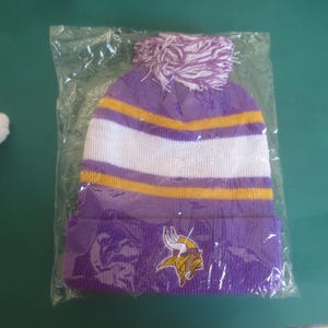 May include: A purple, white, and gold knit beanie with a pom-pom. The hat features a team logo on the front. The hat is packaged in a clear plastic bag.