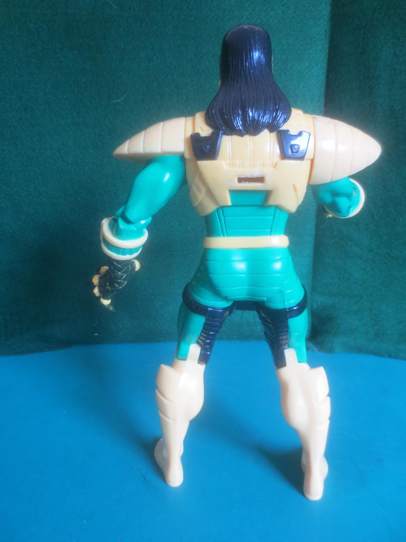 Op de afbeelding: Een plastic actiefiguur van achteren gezien, met lang zwart haar, een cr&egrave;mekleurige borstplaat met zwarte accenten, groenblauwe armen en benen, en cr&egrave;mekleurige laarzen. Het figuur heeft een zwarte handschoen met textuur aan de rechterhand.