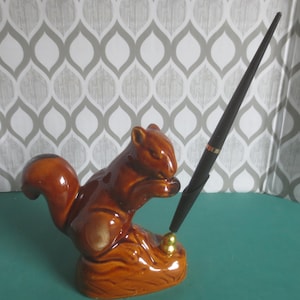 May include: A brown ceramic squirrel pen holder with a black pen. The squirrel is perched on a log-like base. The pen is black with gold accents. The background is a gray and white patterned wallpaper.