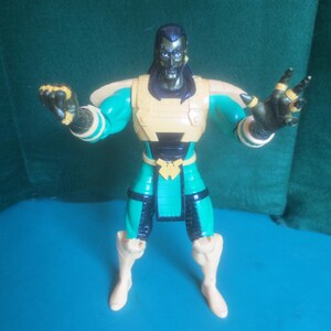 Op de afbeelding: Een plastic actiefiguur met donker haar, een sikje en een metallic groen en beige pak. Het figuur heeft een beige borstplaat, schouderbeschermers en lendendoek, met donkergroene ledematen, zwarte accenten en beige laarzen. Beide armen zijn omhoog.