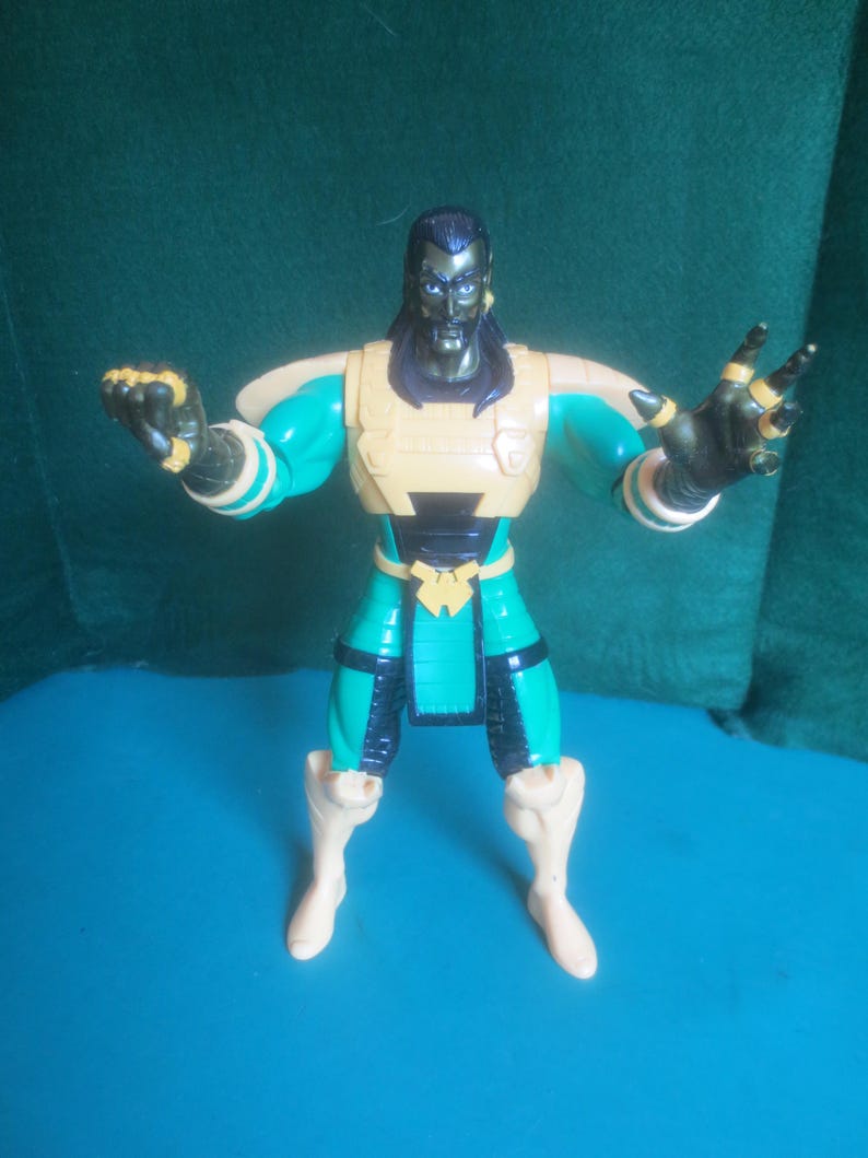 Op de afbeelding: Een plastic actiefiguur met donker haar, een sikje en een metallic groen en beige pak. Het figuur heeft een beige borstplaat, schouderbeschermers en lendendoek, met donkergroene ledematen, zwarte accenten en beige laarzen. Beide armen zijn omhoog.