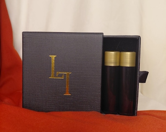 Könnte beinhalten: Eine dunkelgraue Schachtel mit goldfarbenen Buchstaben und zwei zylindrischen Behältern mit goldenen Kappen. Die Schachtel ist geöffnet und zeigt die Behälter im Inneren. Der Hintergrund ist eine Mischung aus weißem und orangefarbenem Stoff.