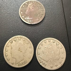 May include: Three antique silver coins. One coin is dated 1908, and the other two are dated 1906 and 1907. Each coin features a profile of a woman and the year of minting. The coins are slightly tarnished, showing signs of age.