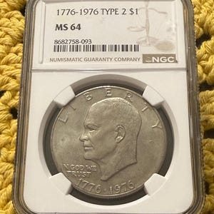 May include: A silver coin from 1776-1976, graded MS 64, in a protective case. The coin shows a man's profile and the inscriptions "IN GOD WE TRUST" and "1776-1976". The case is labelled "NUMISMATIC GUARANTY COMPANY".