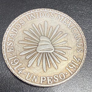 May include: A silver coin from 1914 with the text "ESTADOS UNIDOS MEXICANOS" and "UN PESO" around the edge. The coin features a central design of a radiant sun with a Phrygian cap.