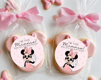 May include: Two clear cellophane bags each containing a Minnie Mouse-themed cookie. Each cookie has a pink frosting base and two pink ear-shaped cookies. The cookies are decorated with a Minnie Mouse graphic and the text "I'M THIS Minnie! KAYLA'S 2ND BIRTHDAY". Pink bow-shaped cookies are scattered around.
