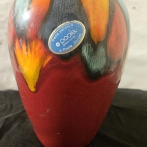 May include: A hand-painted Poole pottery vase with a vibrant color scheme. The vase features a red base that transitions to a blend of orange, yellow, blue, and black hues. A blue and white sticker on the vase reads "Hand painted Poole in Poole, Dorset."