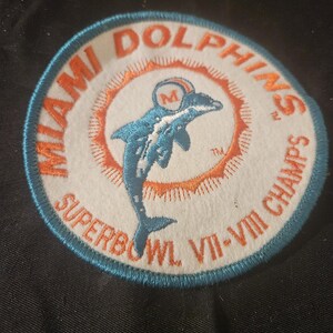 May include: Circular patch featuring the Miami Dolphins logo, a blue dolphin leaping through the air. The patch has an orange and white design with the words "MIAMI DOLPHINS" and "SUPERBOWL VII-VIII CHAMPS" in orange and blue.