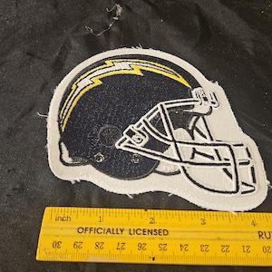 May include: A navy blue and yellow embroidered patch of a football helmet. The helmet has a white outline and a yellow lightning bolt design. The patch is approximately 3 inches in length. The text "OFFICIALLY LICENSED" is visible.