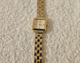 May include: A gold and silver-toned Gruen quartz watch with a square face. The watch features a white dial with gold hands and markers, and a band with alternating gold and silver links. The watch is adorned with small crystals around the face.