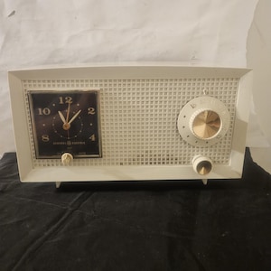 May include: White vintage General Electric clock radio with a black clock face and gold knobs. The radio has a white grid pattern on the front.