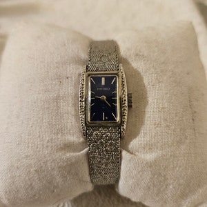 May include: A silver watch with a blue face and a mesh band. The watch face is rectangular and has a silver border. The watch is on a white surface.