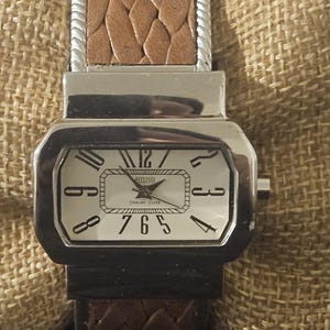 May include: A rectangular silver watch with a white face and black numerals. The watch has a brown leather band with a woven texture. The watch face displays the brand name "Ecclissi".