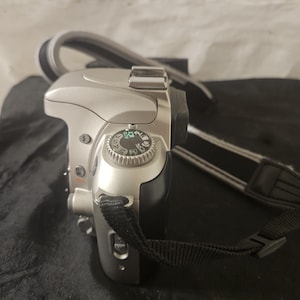 May include: Silver digital camera with a black and gray strap. The camera has a mode dial on top and a black grip. The strap is attached to the camera body. The camera is on a black surface.