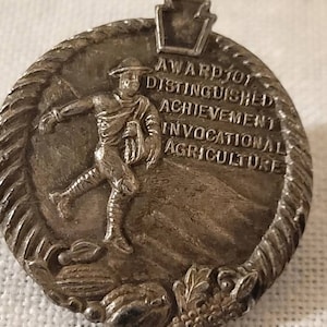 May include: A silver-toned metal pin with a raised image of a person holding a sheaf of wheat. The pin features the text "Award for Distinguished Achievement Vocational Agriculture" and a stylized image of the state of Pennsylvania.