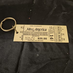 May include: A vintage Miss America pageant ticket keychain from 1991. The ticket is beige with black text, including "Miss America" and "Atlantic City Convention Center." The keychain has a gold-colored ring.