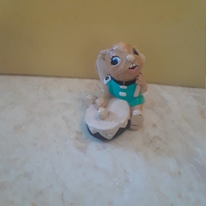 May include: A figurine of a brown rabbit wearing a teal shirt and playing a drum. The rabbit has large blue eyes and is sitting on a drum with a black rim. The figurine is on a white and tan surface.