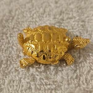May include: A gold-colored turtle-shaped brooch. The brooch has a textured shell and detailed flippers, head, and tail. The turtle is resting on a white, textured surface. The brooch is likely made of metal and is a decorative accessory.