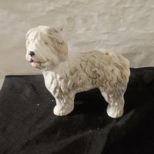 May include: A ceramic figurine of a shaggy dog with a white coat featuring greyish-brown patches on its head and back. The dog is depicted in a standing position with a black nose, visible tongue, and textured fur detailing across its entire body.