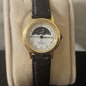 May include: Gold-tone Armitron Quartz watch with a white face featuring a moon and stars design. The watch has a black leather band and a gold bezel. The watch is displayed on a beige velvet surface.
