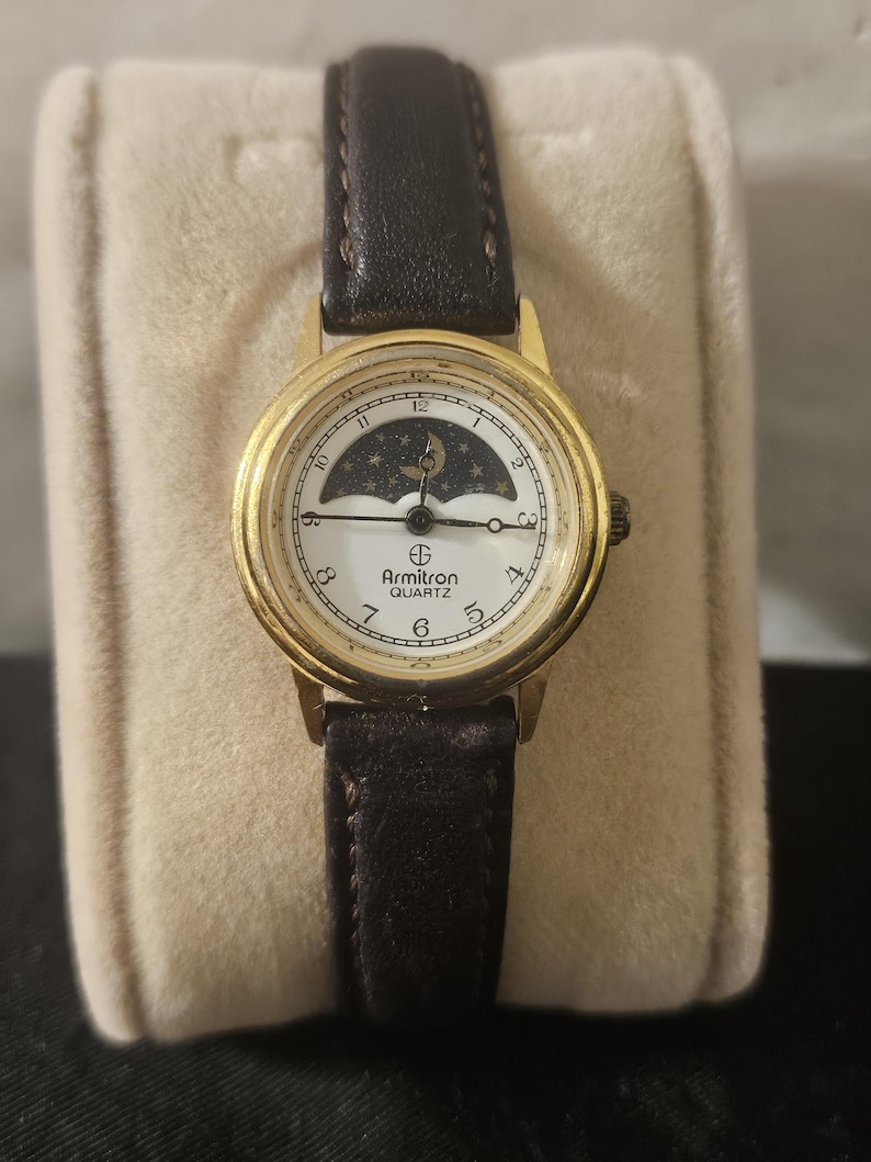 May include: Gold-tone Armitron Quartz watch with a white face featuring a moon and stars design. The watch has a black leather band and a gold bezel. The watch is displayed on a beige velvet surface.