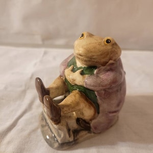 May include: A ceramic figurine of a frog wearing a purple coat and green pants, sitting on a stone base. The frog has yellow eyes and a brown nose.