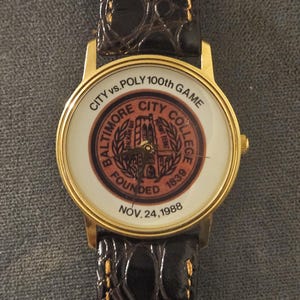 May include: A gold-toned wristwatch with a brown leather band. The watch face features the words "CITY vs. POLY 100th GAME" and "BALTIMORE CITY COLLEGE" in a circular design. The watch was founded in 1839 and the date Nov. 24, 1988 is also displayed.