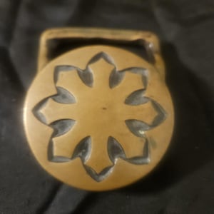 May include: A brass-colored belt buckle with a circular design. The buckle features a central floral pattern with six symmetrical petals. The rectangular loop is attached to the top of the circular design.